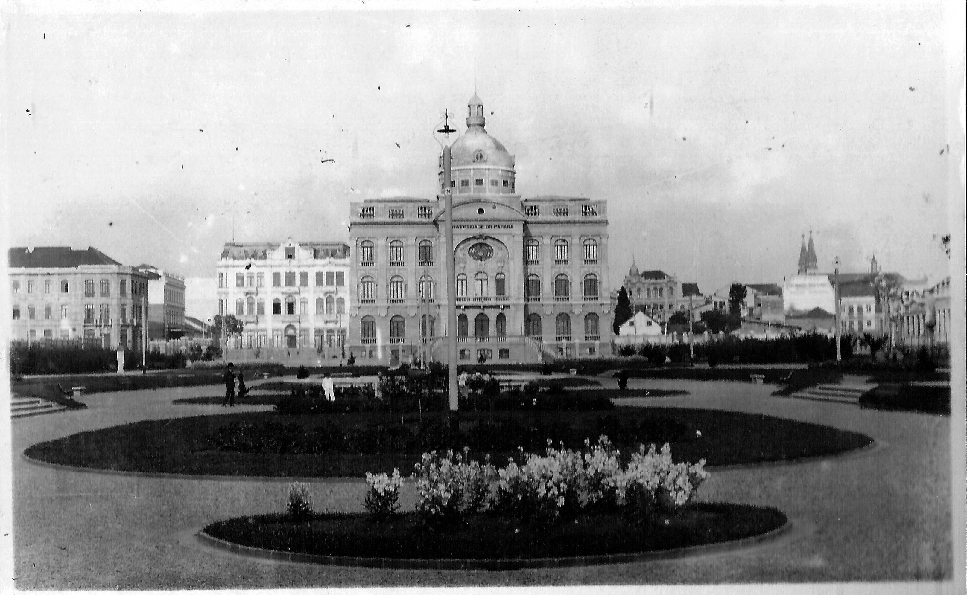 Postal da Praça Santos Andrade no início da década de 1920.  O espaço apresenta as intervenções de 1917, com paisagismo à francesa, apresentando jardins e passeios simetricamente organizados. Ao fundo, o prédio da Universidade do Paraná, com a imponente cúpula, demolida na década de 1950.